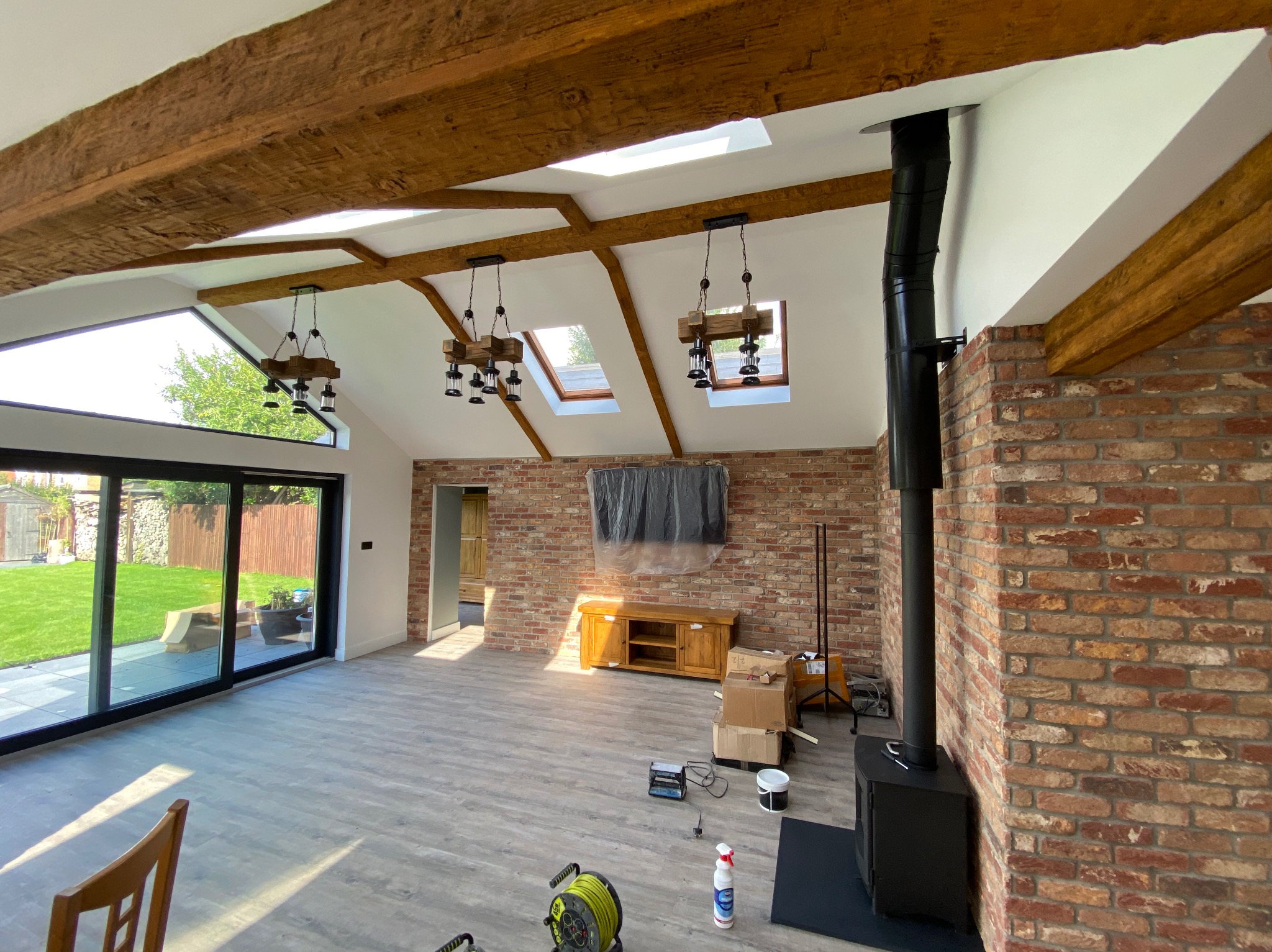 Przestronne wnętrze po remoncie: ceglana ściana z kominem, drewniane belki stropowe, duże okna z widokiem na ogród, drewniane lampy. Podłoga wyłożona panelami.