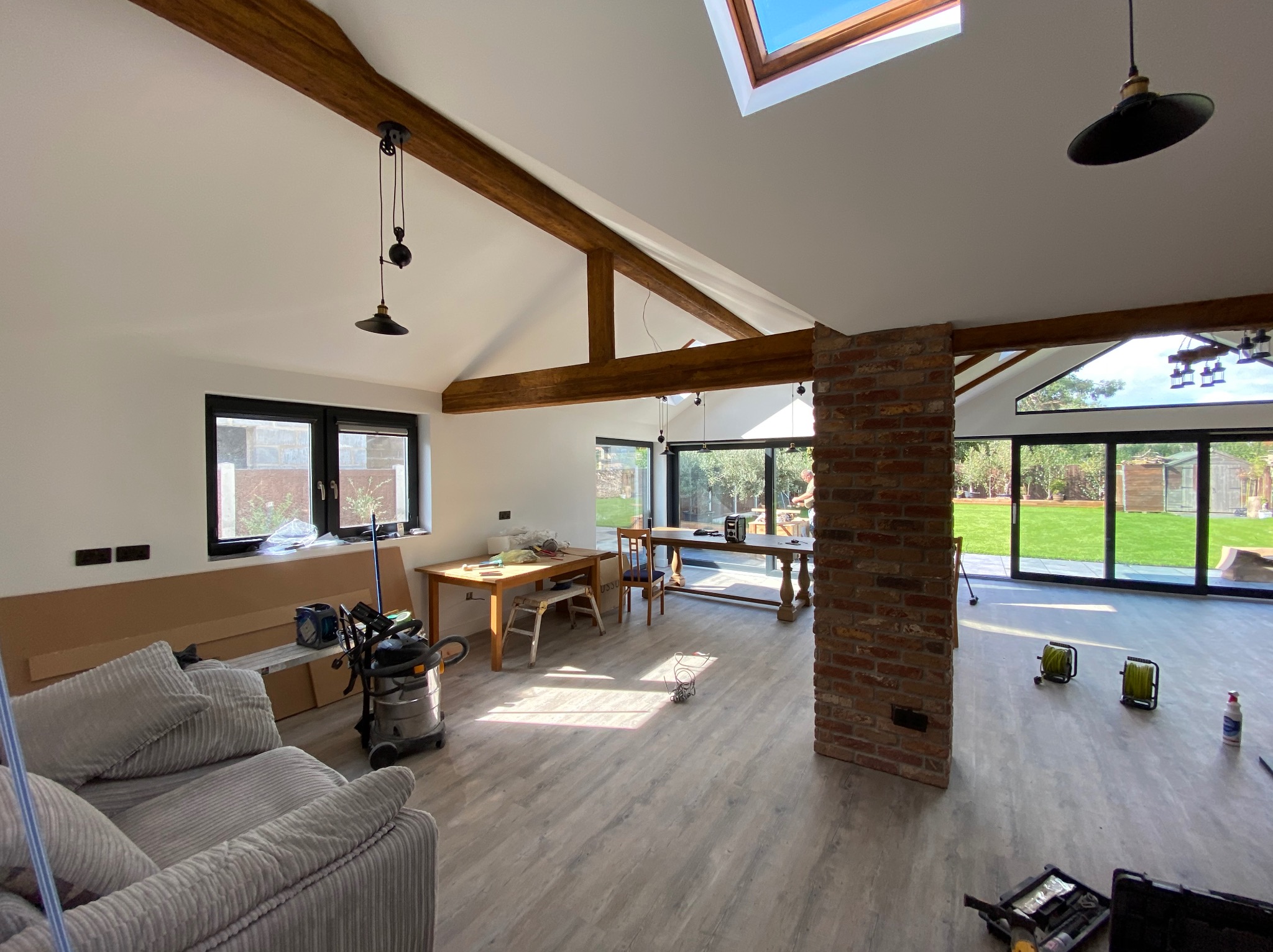 Jasne wnętrze po remoncie z drewnianymi belkami stropowymi, dużymi oknami i podłogą w odcieniach szarości. Widoczny fragment sofy i narzędzia.