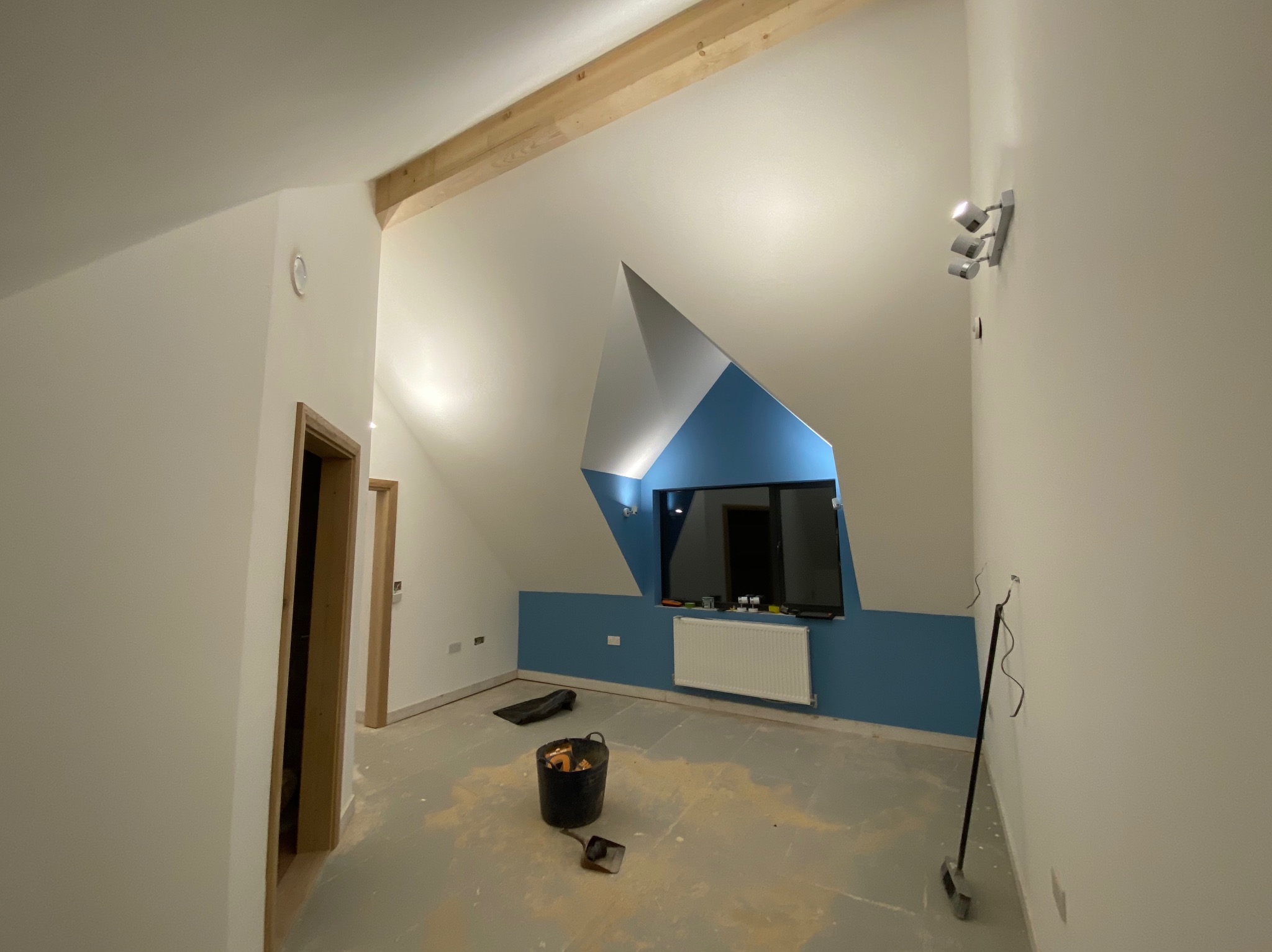 Pokój na poddaszu w trakcie wykańczania: pomalowane ściany na biało i niebiesko, drewniana belka, okno, grzejnik, widoczne narzędzia i wiadro z farbą.