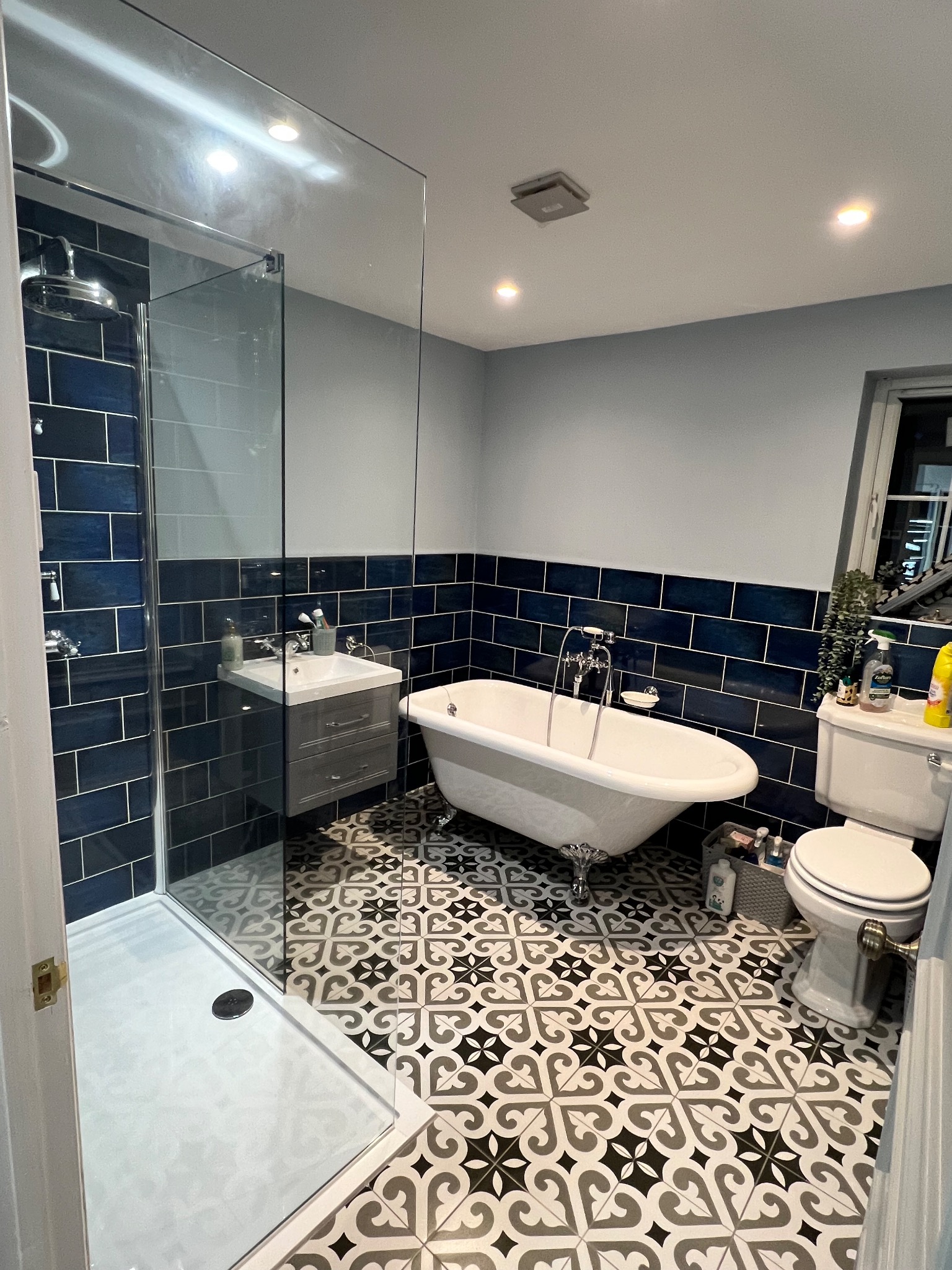 Wykończona łazienka z kabiną prysznicową, wolnostojącą wanną na nóżkach i wzorzystą podłogą. Ciemne, błyszczące płytki do połowy ściany. Nowoczesny design.