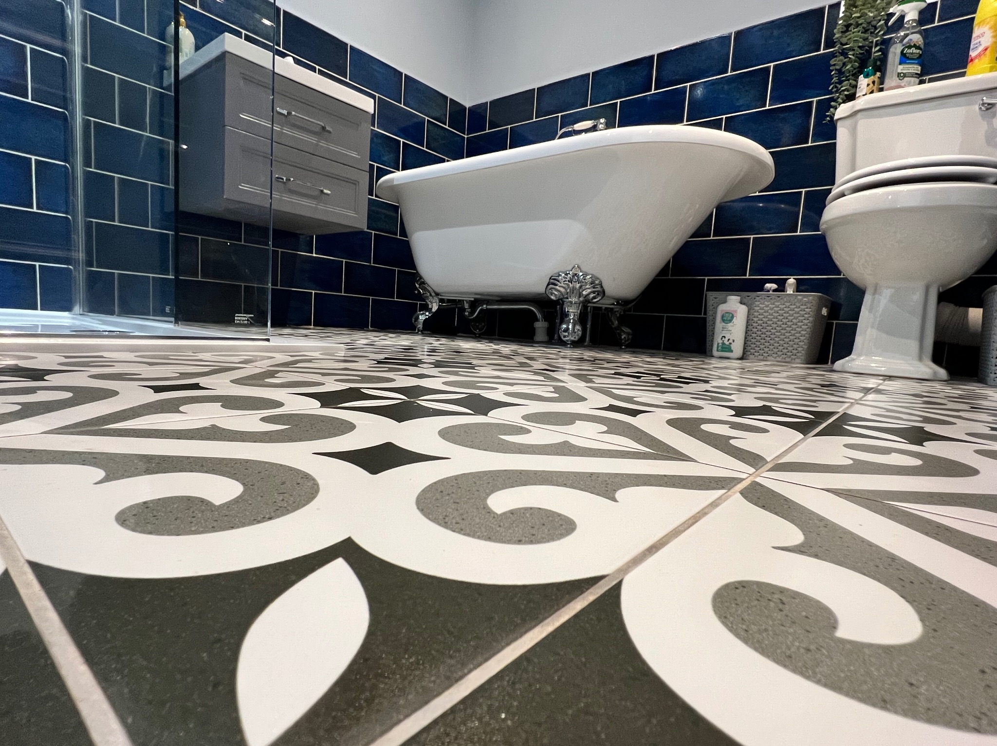 Wykończona łazienka z wolnostojącą wanną na ozdobnych nóżkach, umywalką z szafką i toaletą. Na ścianach ciemnoniebieskie płytki, na podłodze wzorzysta mozaika.