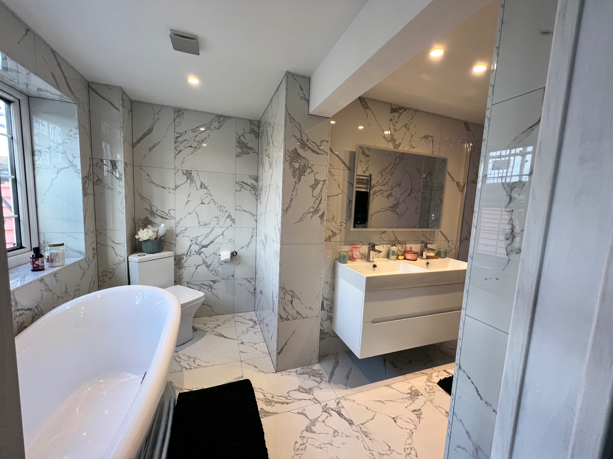 Elegancka łazienka z wanną wolnostojącą i marmurowymi płytkami. Umywalka nablatowa z szafką, lustro. Nowoczesne oświetlenie sufitowe. Czarny dywanik.