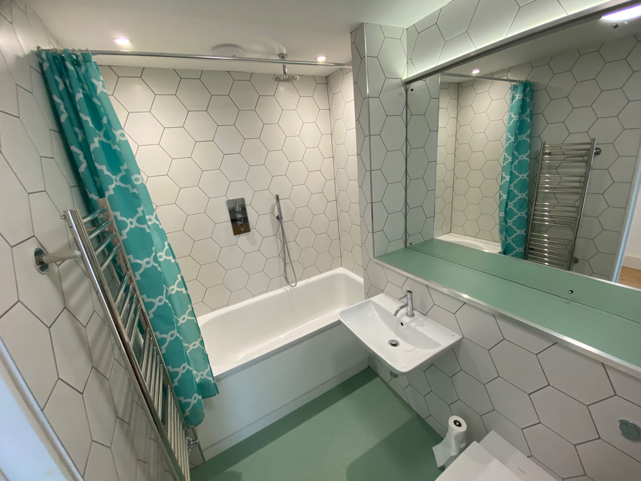 Nowoczesna łazienka z heksagonalnymi płytkami, turkusową zasłoną prysznicową i podłogą w odcieniu mięty. Minimalistyczna umywalka i grzejnik drabinkowy.