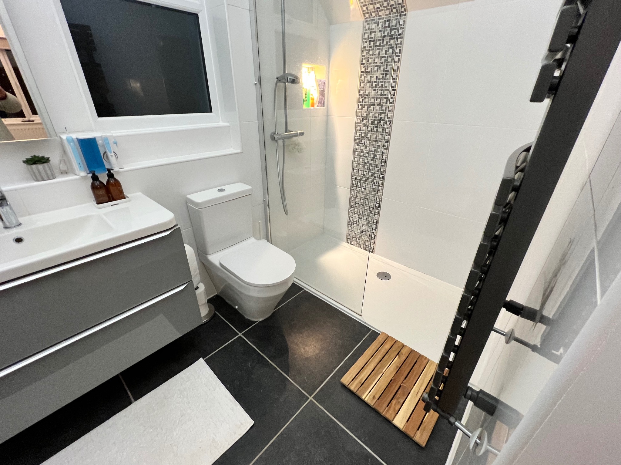 Nowoczesna łazienka po remoncie: szare szafki pod umywalką, biała toaleta, kabina prysznicowa z mozaiką i czarne płytki podłogowe z drewnianym dywanikiem.