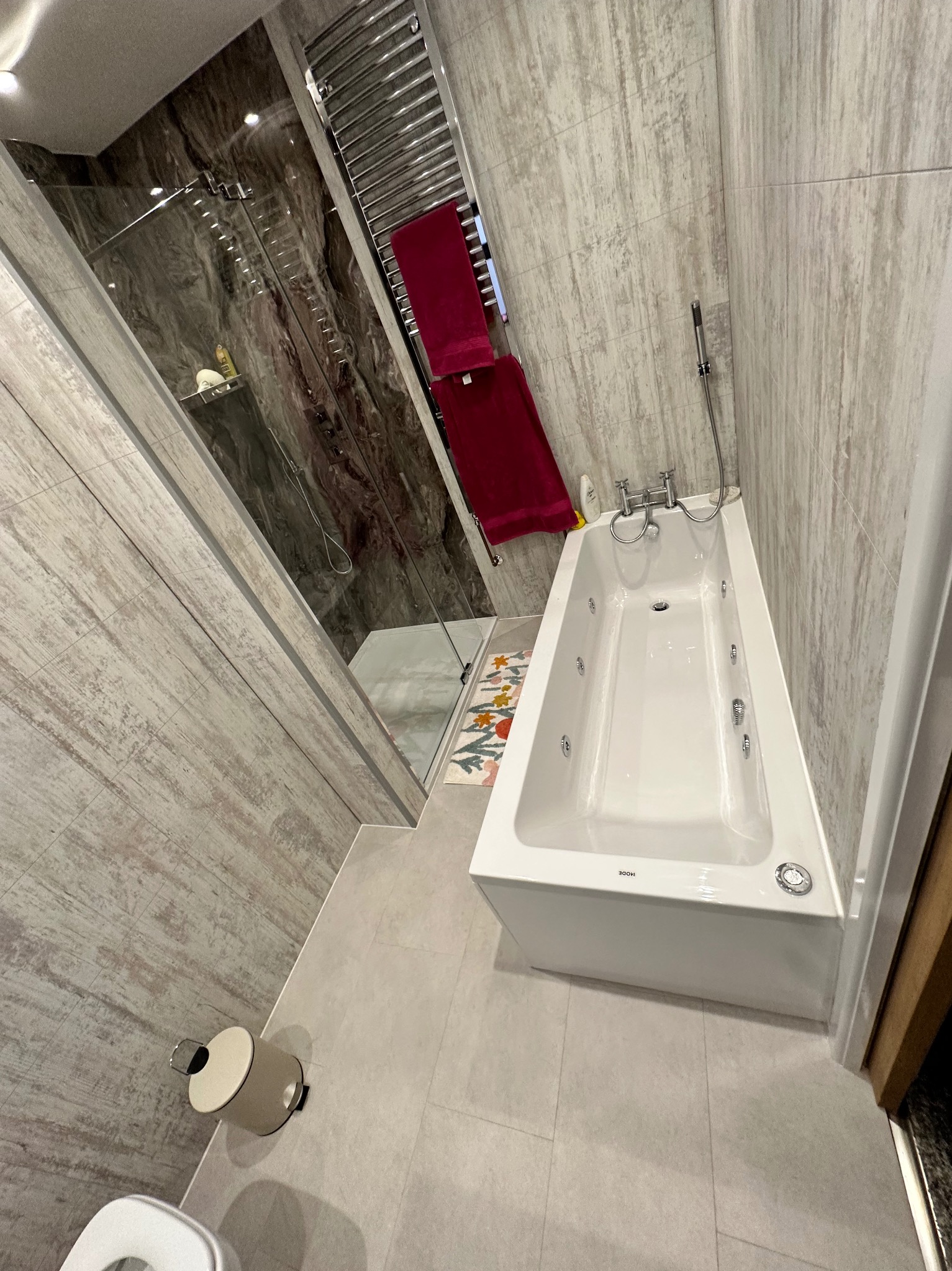 Nowoczesna łazienka po remoncie: biała wanna z hydromasażem, szklana kabina prysznicowa, grzejnik drabinkowy z czerwonymi ręcznikami, płytki imitujące drewno.