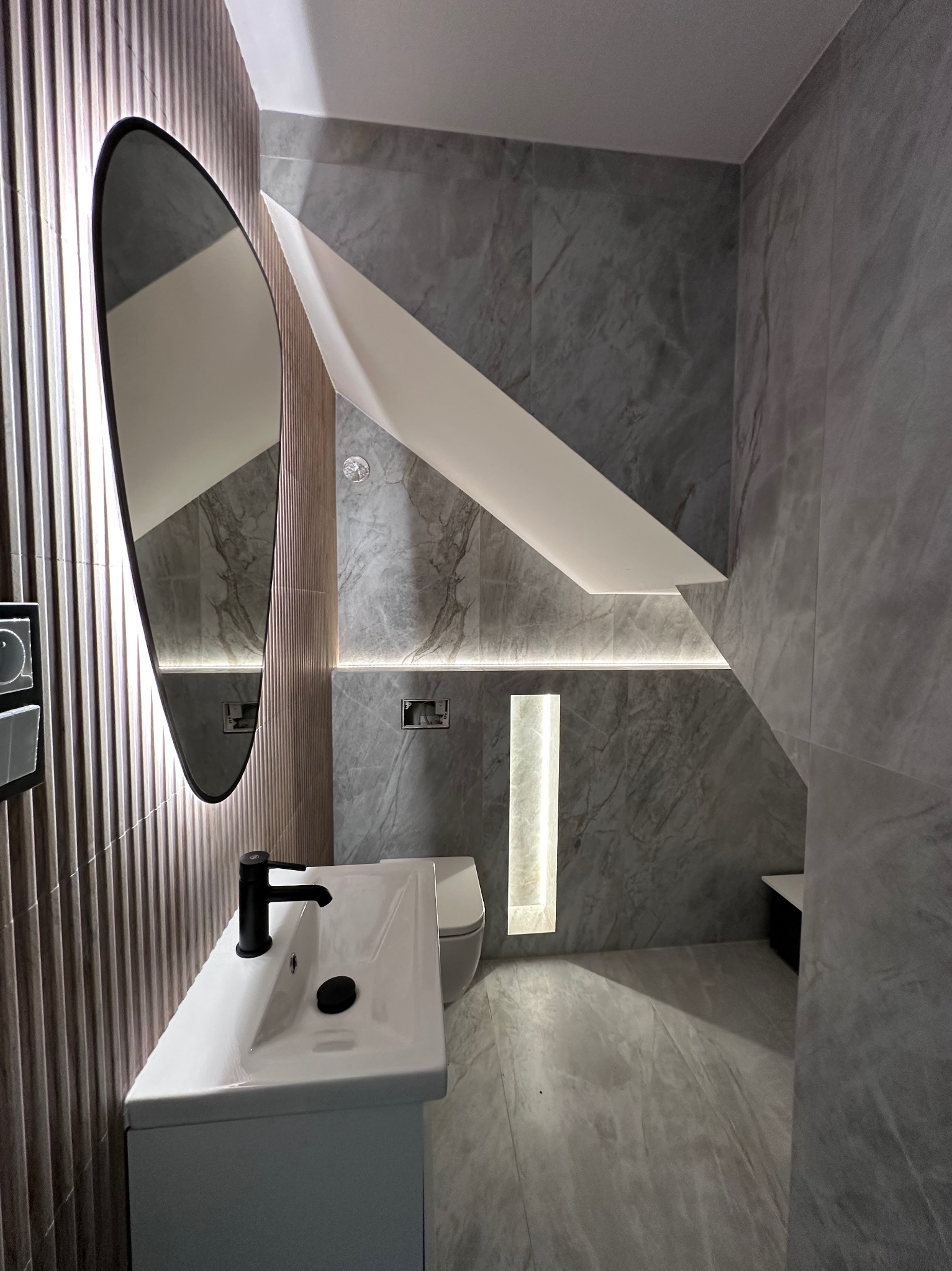 Nowoczesna łazienka w szarościach z podświetleniem LED. Umywalka z czarną armaturą, owalne lustro i marmurowe płytki tworzą elegancki wystrój.