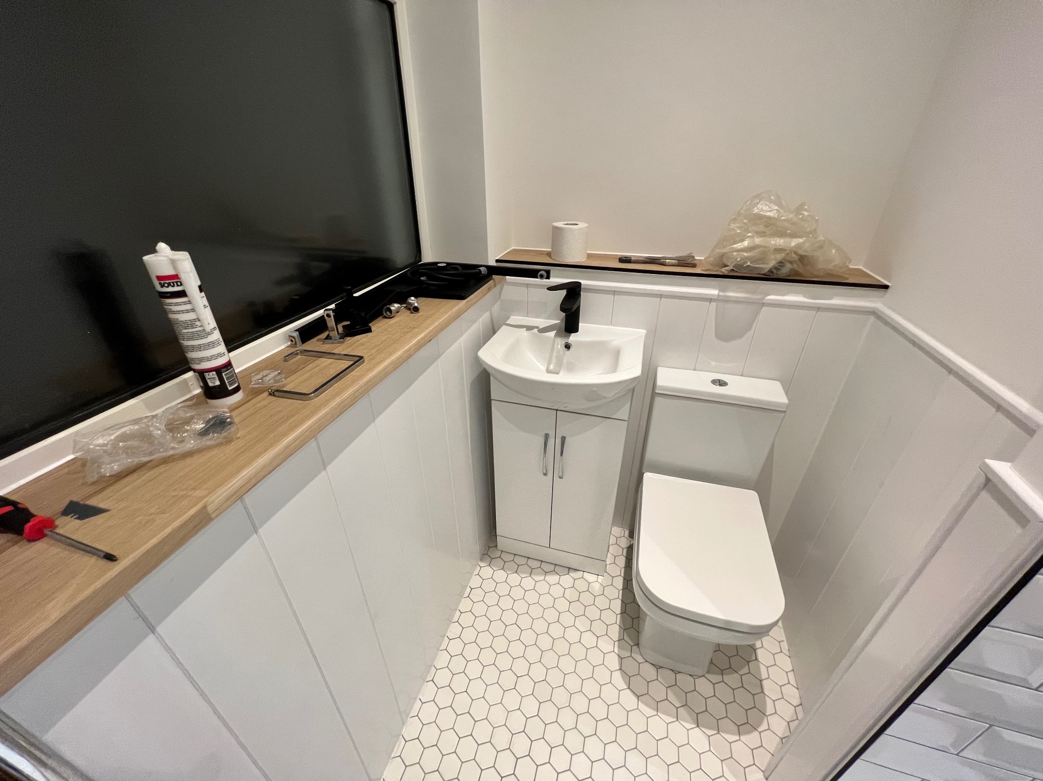 Biała, wąska umywalka z szafką i toaleta w małej łazience po remoncie. Na blacie silikon, narzędzia i akcesoria. Heksagonalna podłoga i boazeria.