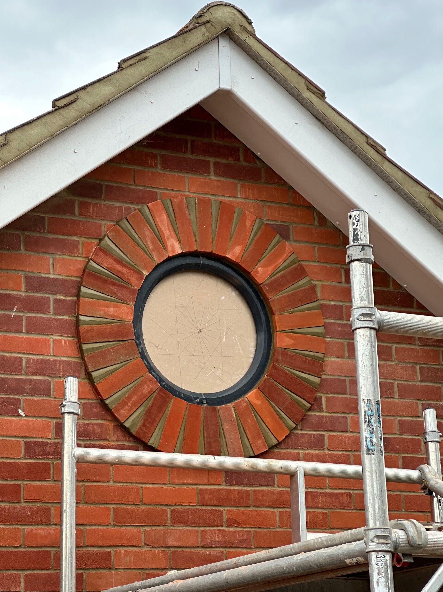 Oryginalne okrągłe okno z ceglaną obudową w trakcie montażu na czerwonej ścianie z cegieł. Widoczne rusztowanie. Nietypowy detal architektoniczny.