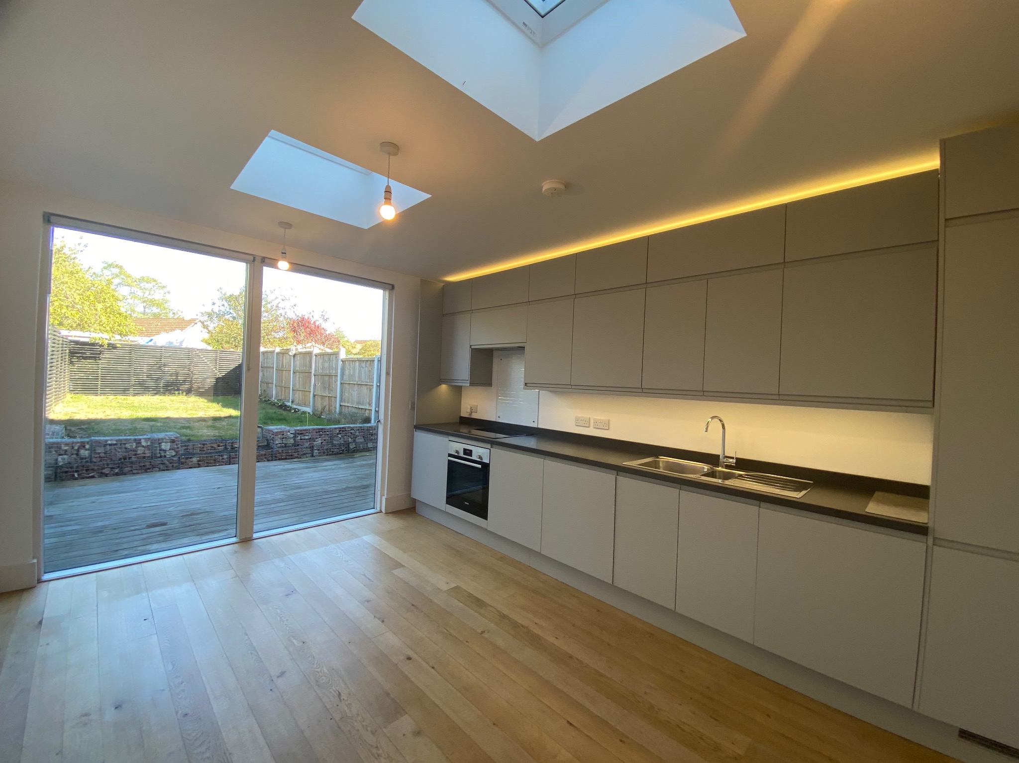 Jasna kuchnia z drewnianą podłogą i dużym oknem na ogród. Minimalistyczne szafki z oświetleniem LED. Dwa okna dachowe wpuszczają naturalne światło.