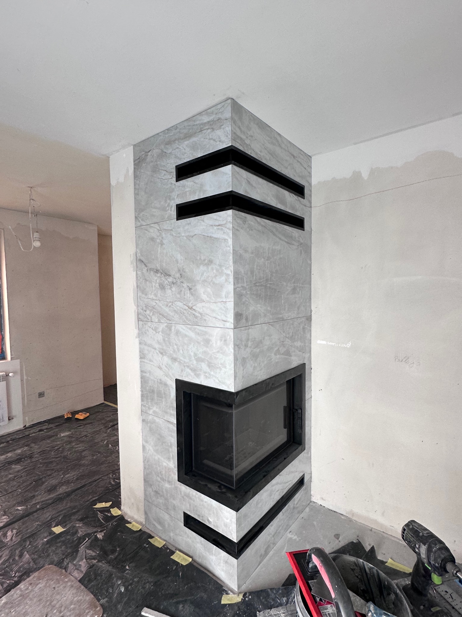 Obudowa kominka z szarych płyt imitujących marmur, z czarnymi wstawkami, w trakcie prac wykończeniowych. Podłoga zabezpieczona folią, widoczne narzędzia.