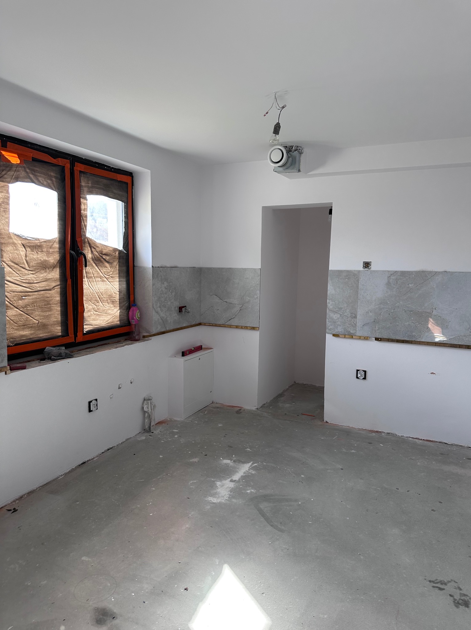 Wnętrze w trakcie prac: szare płytki na ścianie, surowa podłoga, okno z folią ochronną. Widoczne instalacje elektryczne i poziomica. Etap przygotowania do dalszych prac.