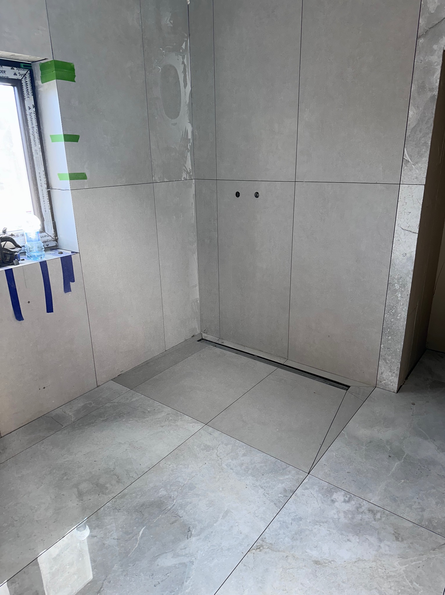 Wykończona łazienka z szarymi płytkami, widoczny odpływ liniowy i przygotowane punkty pod armaturę. Okno zabezpieczone taśmą malarską.