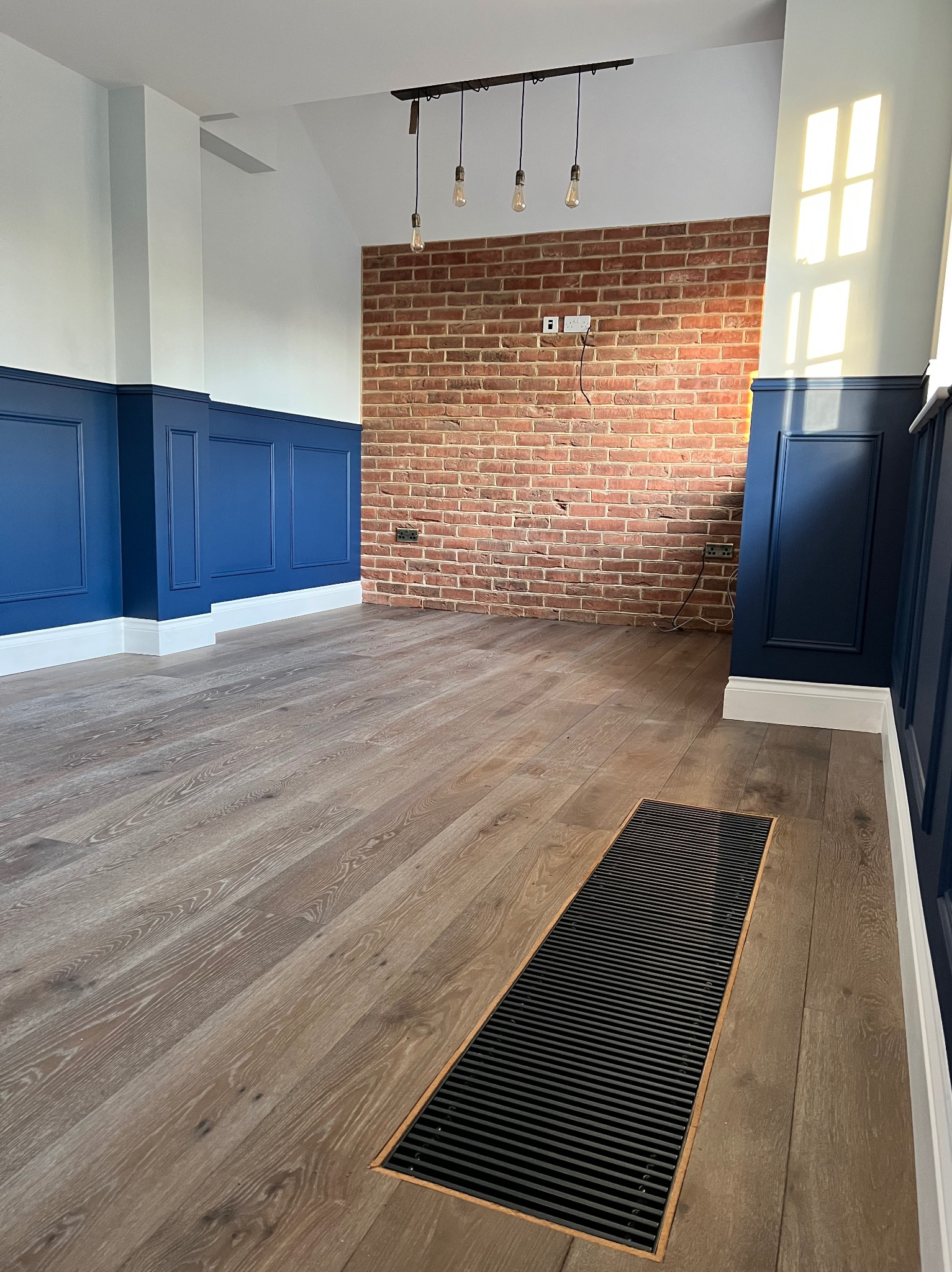 Wnętrze po renowacji: panele ścienne w odcieniach błękitu, ceglana ściana z gniazdkami, drewniana podłoga z kratką wentylacyjną i wiszące lampy. Nowoczesny design.