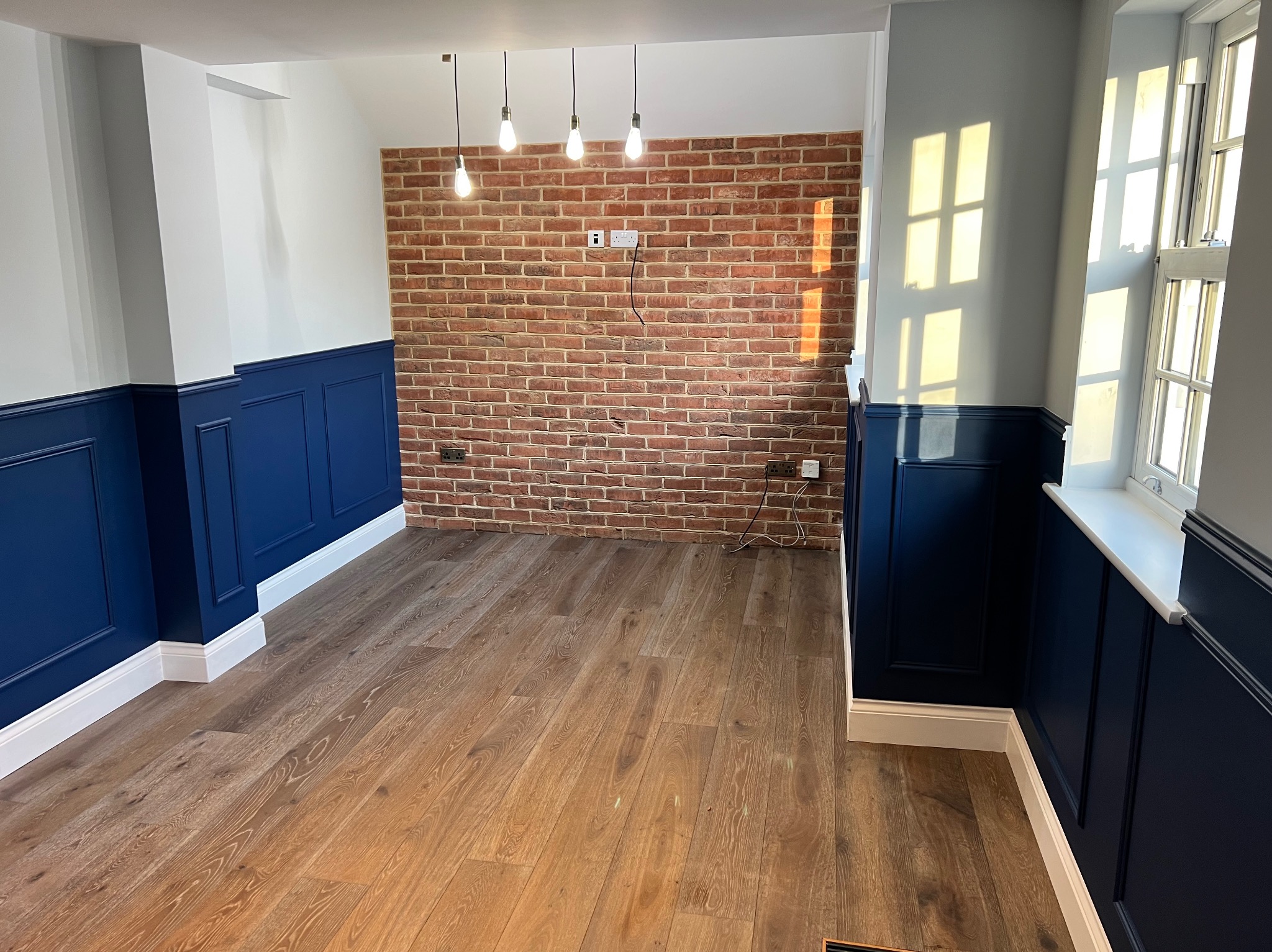 Eleganckie wykończenie wnętrza: drewniana podłoga, ściany z cegły i granatowa boazeria. Trzy żarówki wiszą na tle ceglanej ściany. Promienie słońca na ścianie.