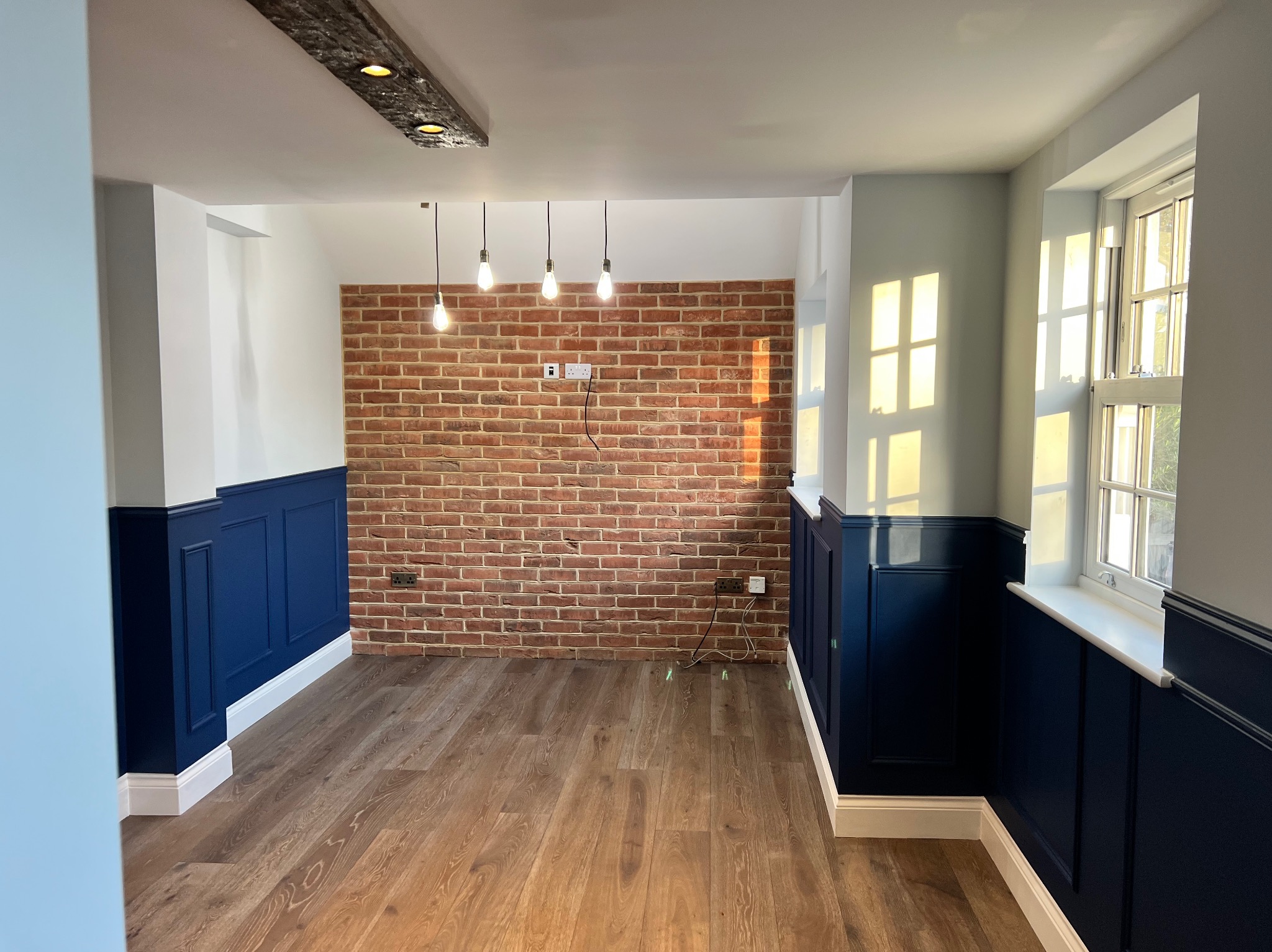 Eleganckie wnętrze z drewnianą podłogą, ceglaną ścianą i granatową boazerią. Wiszące żarówki i naturalne światło wpadające przez okno tworzą przytulny klimat.