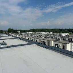 Marcin Czapla - Widok z góry na płaskie dachy szeregu domów w Świętochłowicach, z widocznymi kominami i obróbkami blacharskimi. W tle zieleń i błękitne niebo.