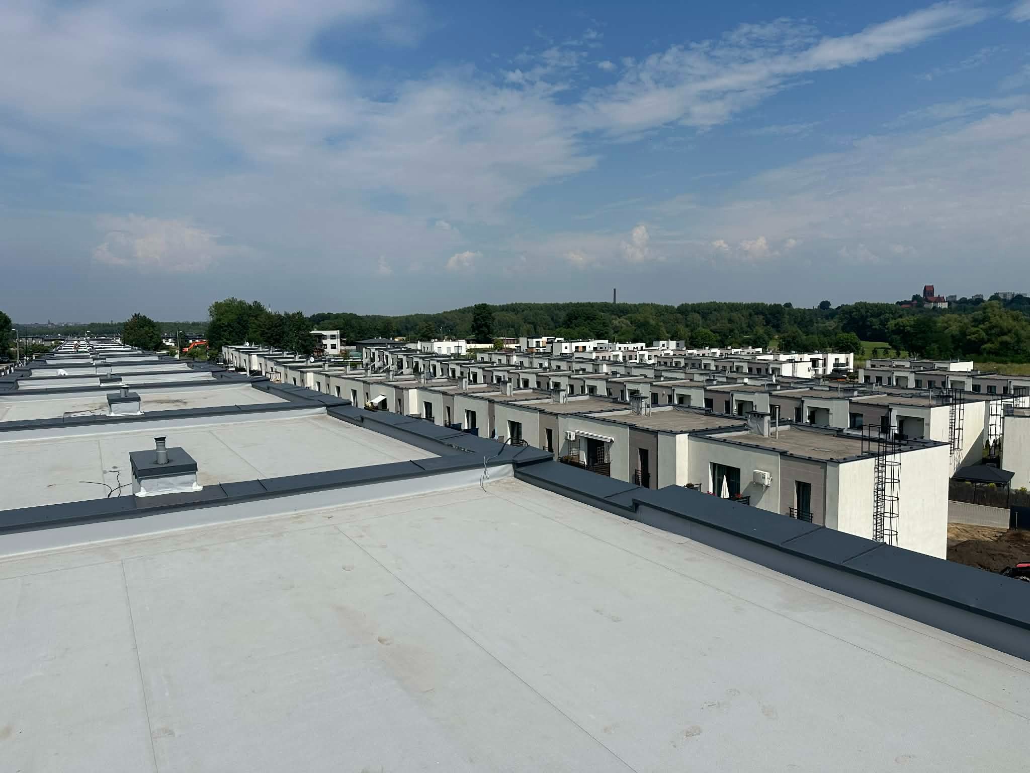 Widok z góry na płaskie dachy szeregu domów w Świętochłowicach, z widocznymi kominami i obróbkami blacharskimi. W tle zieleń i błękitne niebo.