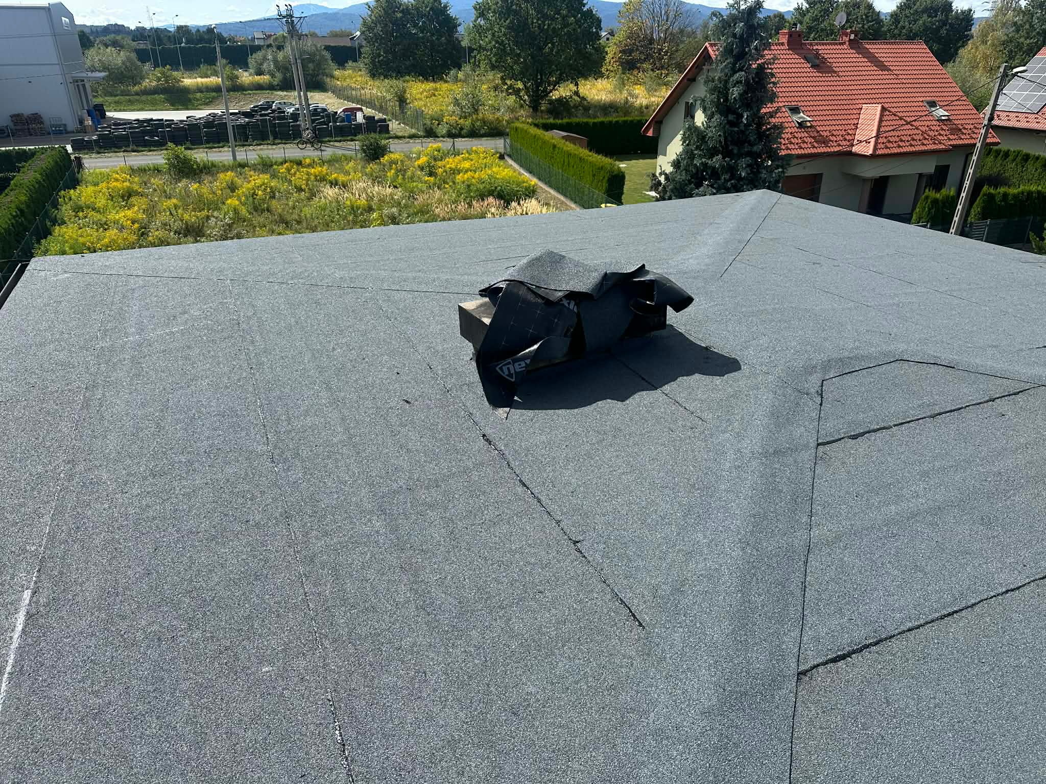 Szary, płaski dach pokryty papą termozgrzewalną z widocznymi łączeniami. Na dachu leżą resztki papy. W tle zabudowa jednorodzinna i zieleń.