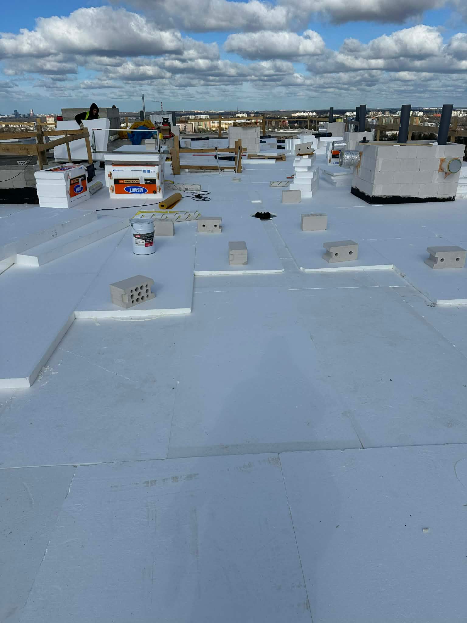 Ocieplenie dachu styropianem na płaskiej powierzchni. Widoczne bloczki i puszka farby. W tle zabudowa miejska i błękitne niebo z chmurami.