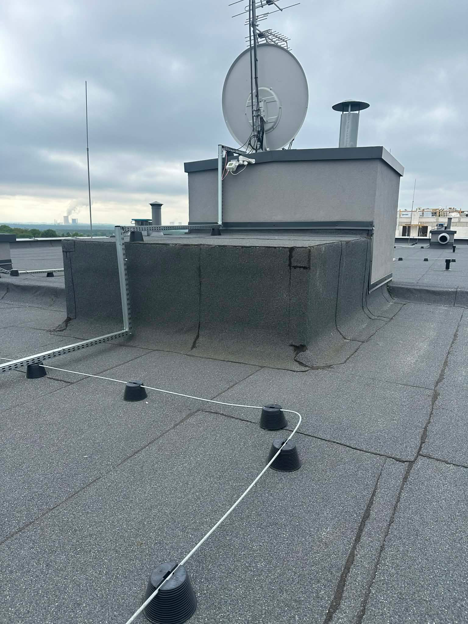 Szary dach pokryty papą z instalacją odgromową, anteną satelitarną i kominem. Widoczne elementy mocujące instalację. W tle pochmurne niebo i kominy elektrowni.