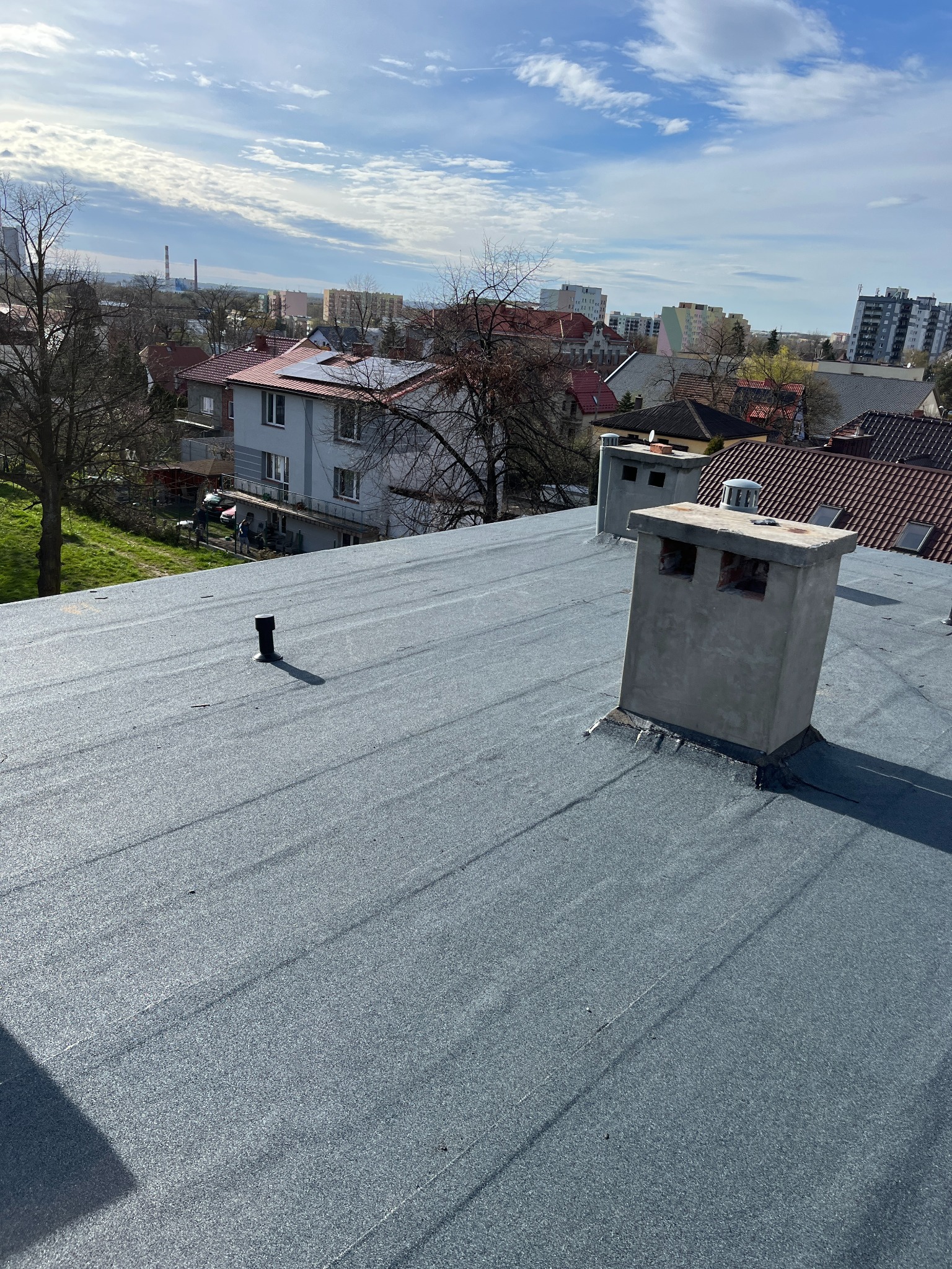 Szary, płaski dach pokryty papą termozgrzewalną z dwoma kominami na tle zabudowy miejskiej i błękitnego nieba z chmurami. Widok z góry, słoneczny dzień.