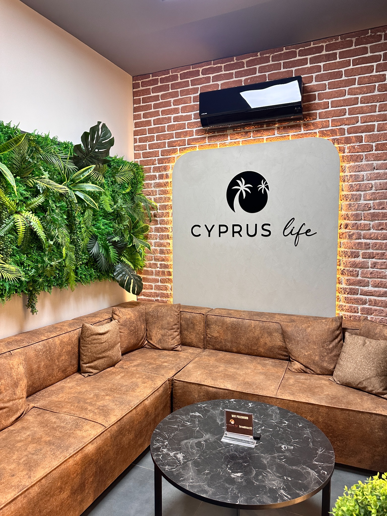 Stylowe wnętrze z narożnikiem w odcieniach brązu, ścianą z cegły i zieloną ścianą z roślin. Na ścianie logo 'Cyprus Life'. Klimatyzator na ścianie.