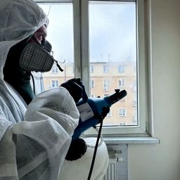 NOWE Goryzonty - Pracownik w kombinezonie ochronnym, masce i rękawicach, rozpyla preparat dezynfekujący w pomieszczeniu, widoczne zaparowane okno i budynek w tle.