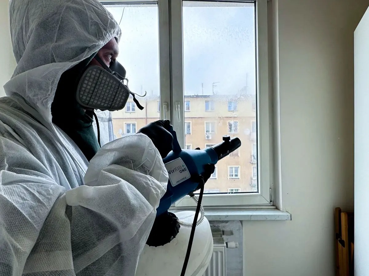 Pracownik w kombinezonie ochronnym, masce i rękawicach, rozpyla preparat dezynfekujący w pomieszczeniu, widoczne zaparowane okno i budynek w tle.