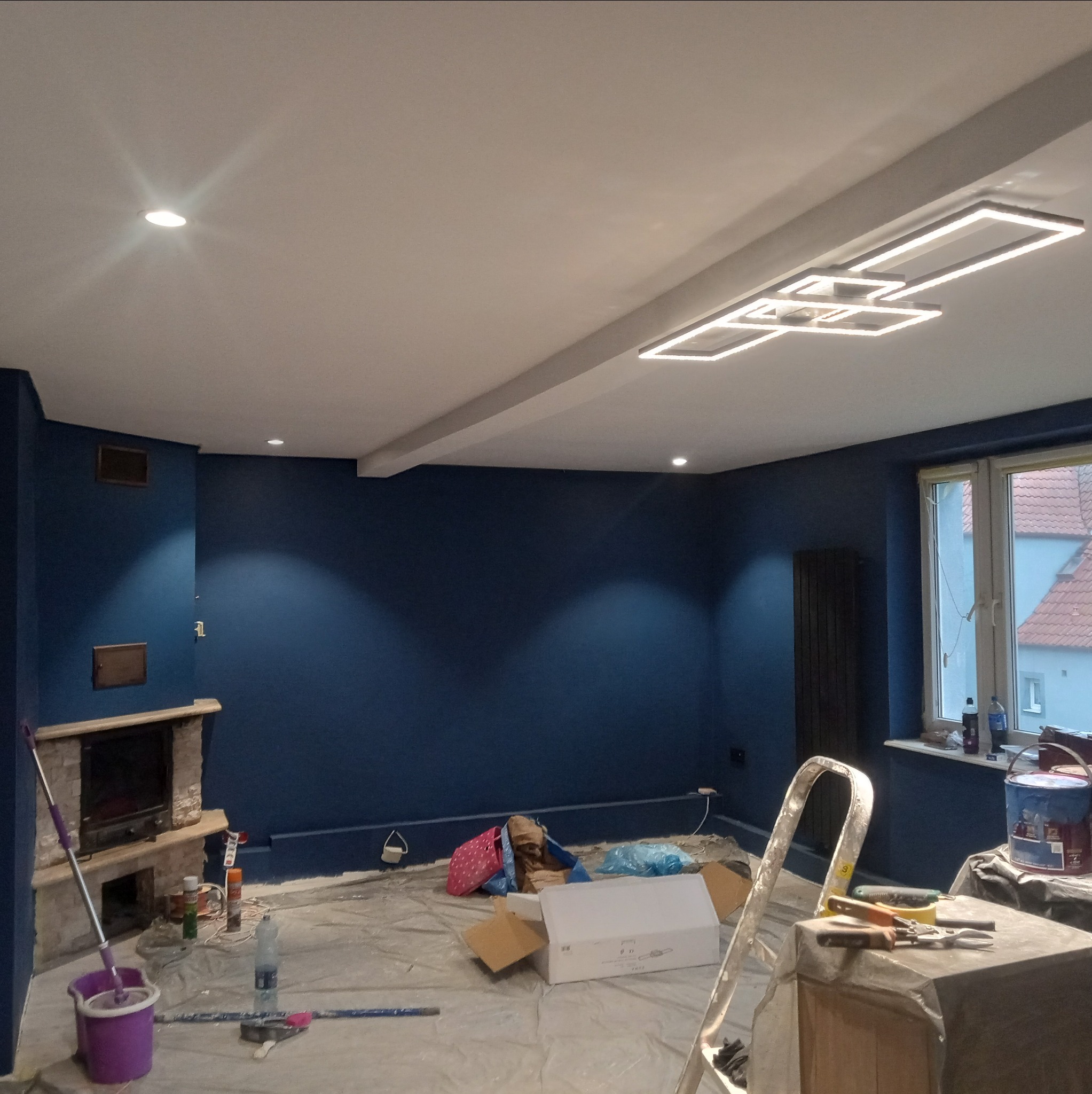 Pokój po malowaniu na granatowo z nowoczesnym oświetleniem sufitowym. Widoczne narzędzia malarskie i zabezpieczenia podłogi. Świeżo odnowione wnętrze.