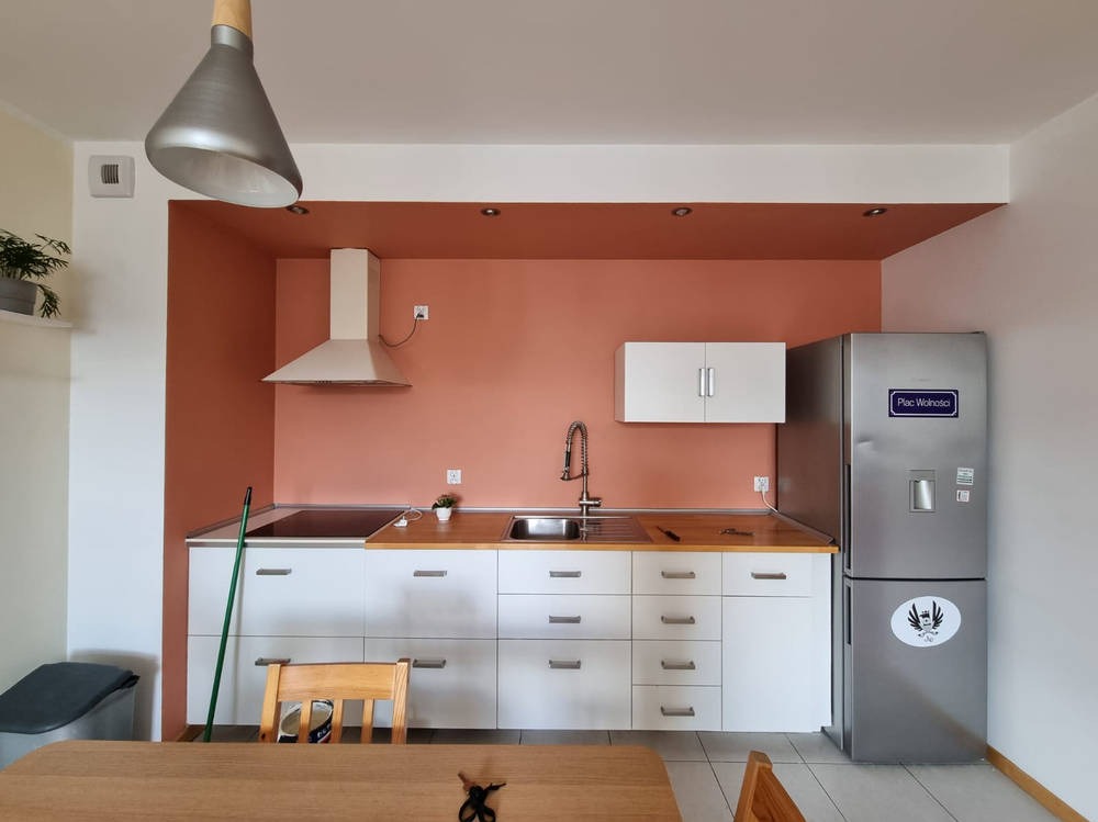 Nowoczesna kuchnia po remoncie: białe szafki z drewnianym blatem, miedziana ściana, srebrna lodówka z naklejkami. Widok na odnowione wnętrze.