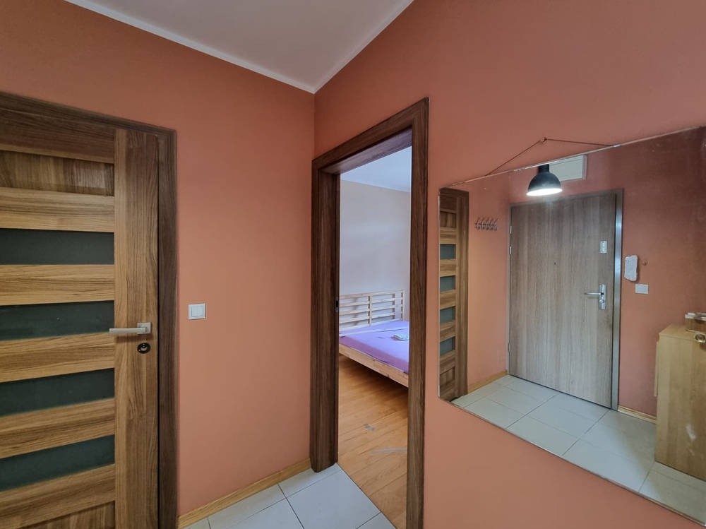 Korytarz po odświeżeniu: pomarańczowe ściany, drewniane drzwi z przeszkleniami, lustro odbijające wejście i fragment sypialni. Nowoczesny design.
