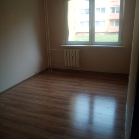 Pusty pokój po remoncie z panelami podłogowymi, grzejnikiem pod oknem i świeżo pomalowanymi ścianami w jasnym odcieniu. Widok na budynek zza okna.