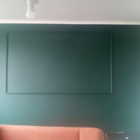 Szmaragdowa ściana z dekoracyjną ramą, oświetlona żarówką. Fragment sofy w odcieniach brzoskwini. Minimalistyczny design wnętrza.