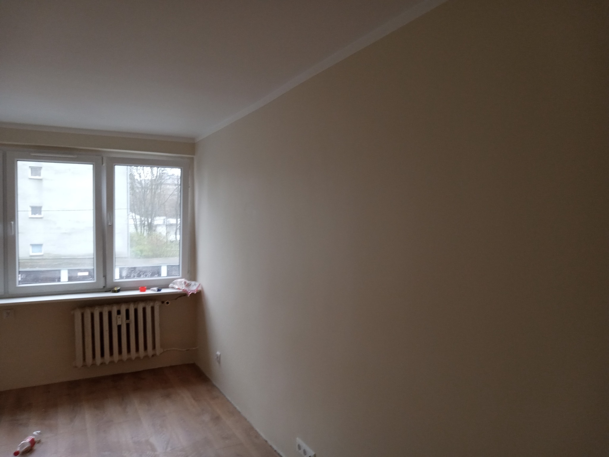 Jasny pokój po malowaniu ścian na beżowo, widoczne okno z grzejnikiem i fragment podłogi. Listwa przypodłogowa i sufitowa w kolorze białym.