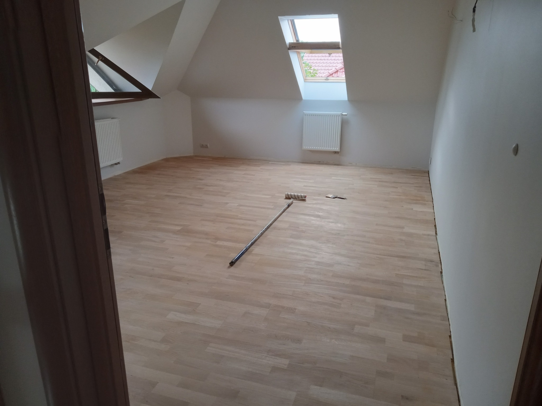 Poddasze po remoncie: jasne panele podłogowe, białe ściany, okno dachowe. Na podłodze wałek malarski i szpachelka. Widok zza framugi drzwi.