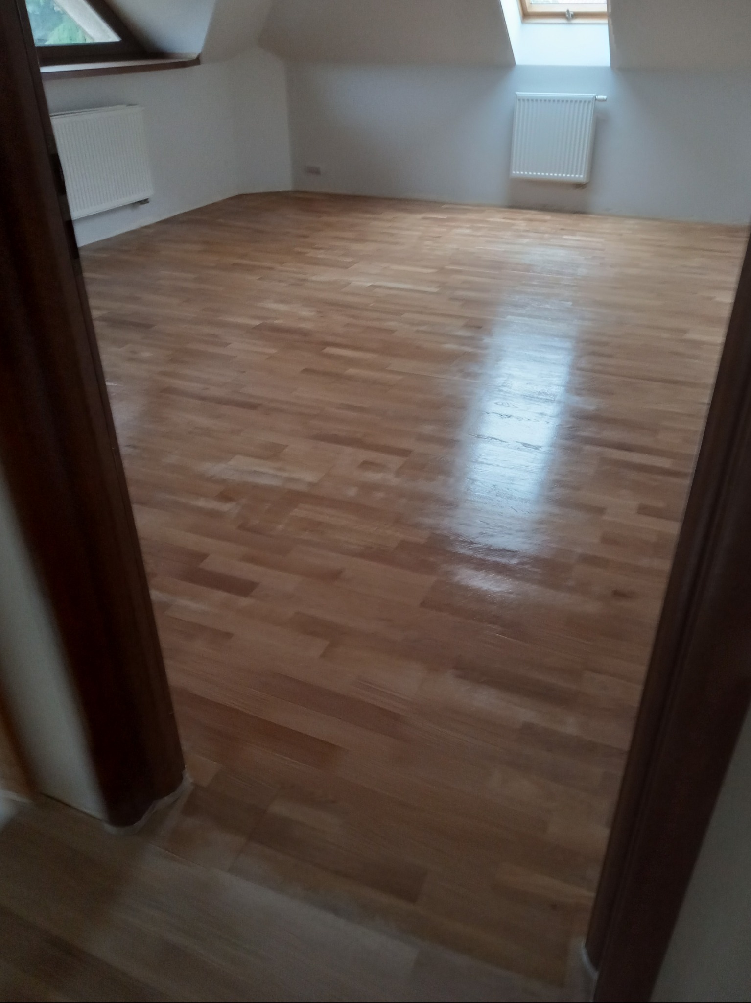 Jasna, drewniana podłoga parkietowa w pokoju na poddaszu z oknami dachowymi i białymi grzejnikami. Widok zza framugi drzwi.