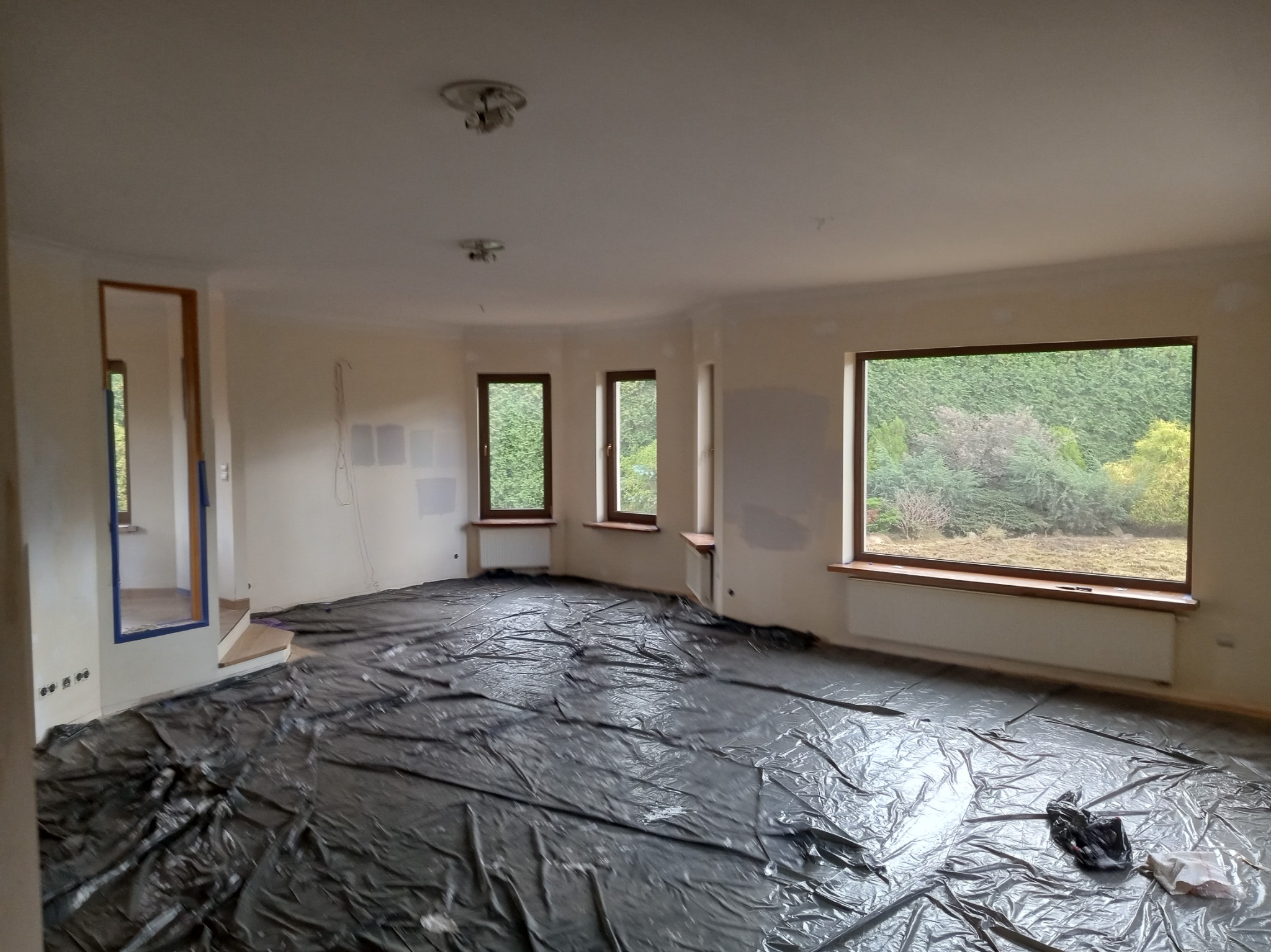 Przygotowanie do malowania: duży pokój z oknami, podłoga zabezpieczona folią malarską, ściany świeżo pomalowane na biało, widoczne ślady po szpachlowaniu.