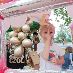 Agnieszka Kalbarczyk Animacja Kids - Uśmiechnięty chłopiec z blond włosami trzyma papierową czapkę błazna na tle dekoracji z balonów i srebrnej konstrukcji. Widoczne osoby w tle.