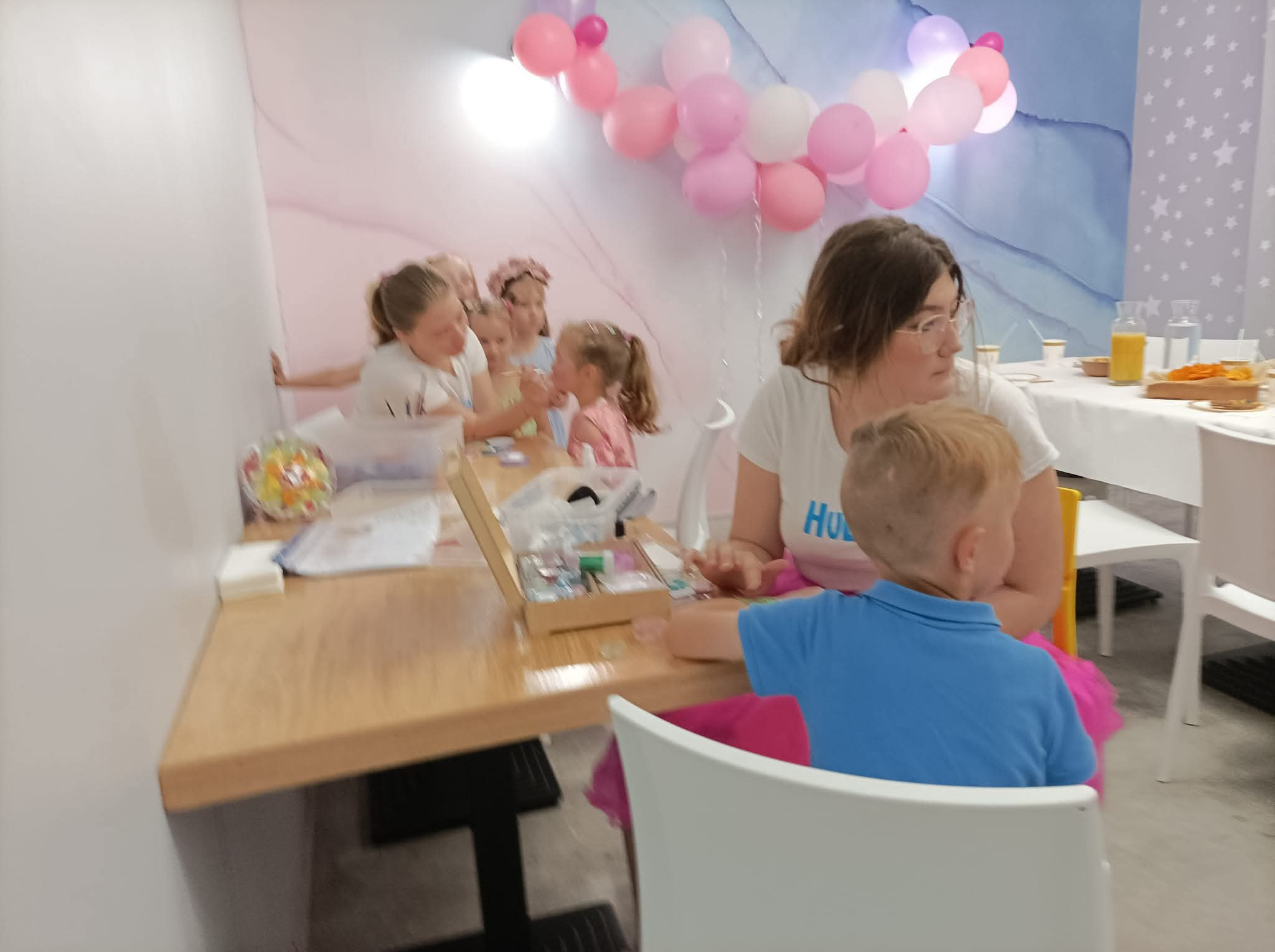 Dzieci przy stole podczas warsztatów plastycznych na tle dekoracji z balonów i wzorzystej ściany. Widoczna kobieta nadzorująca zajęcia i materiały plastyczne.