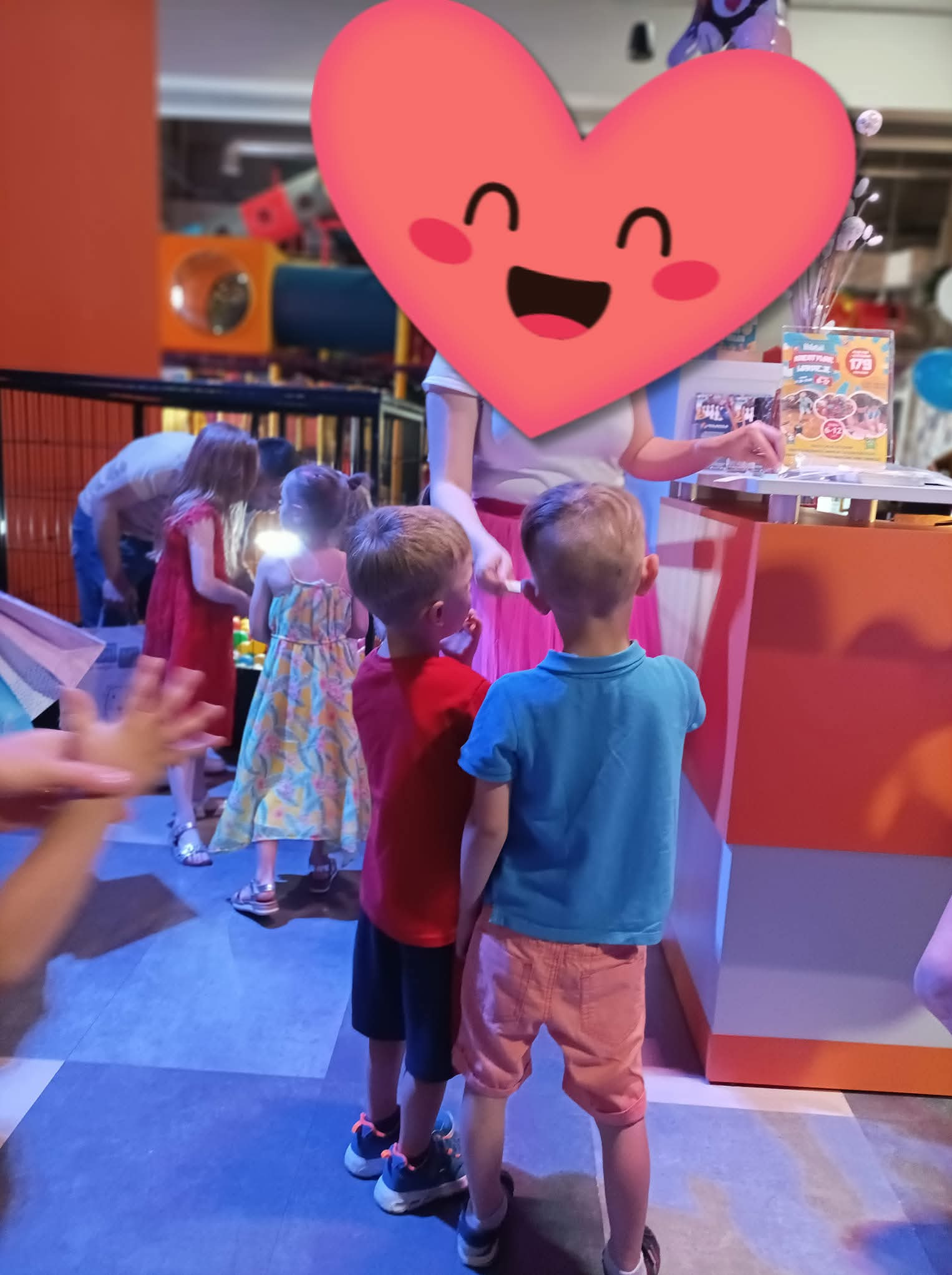 Dzieci w sali zabaw, czekające na animatorkę z zasłoniętą twarzą, która rozdaje upominki. W tle widoczne elementy placu zabaw i inne dzieci.