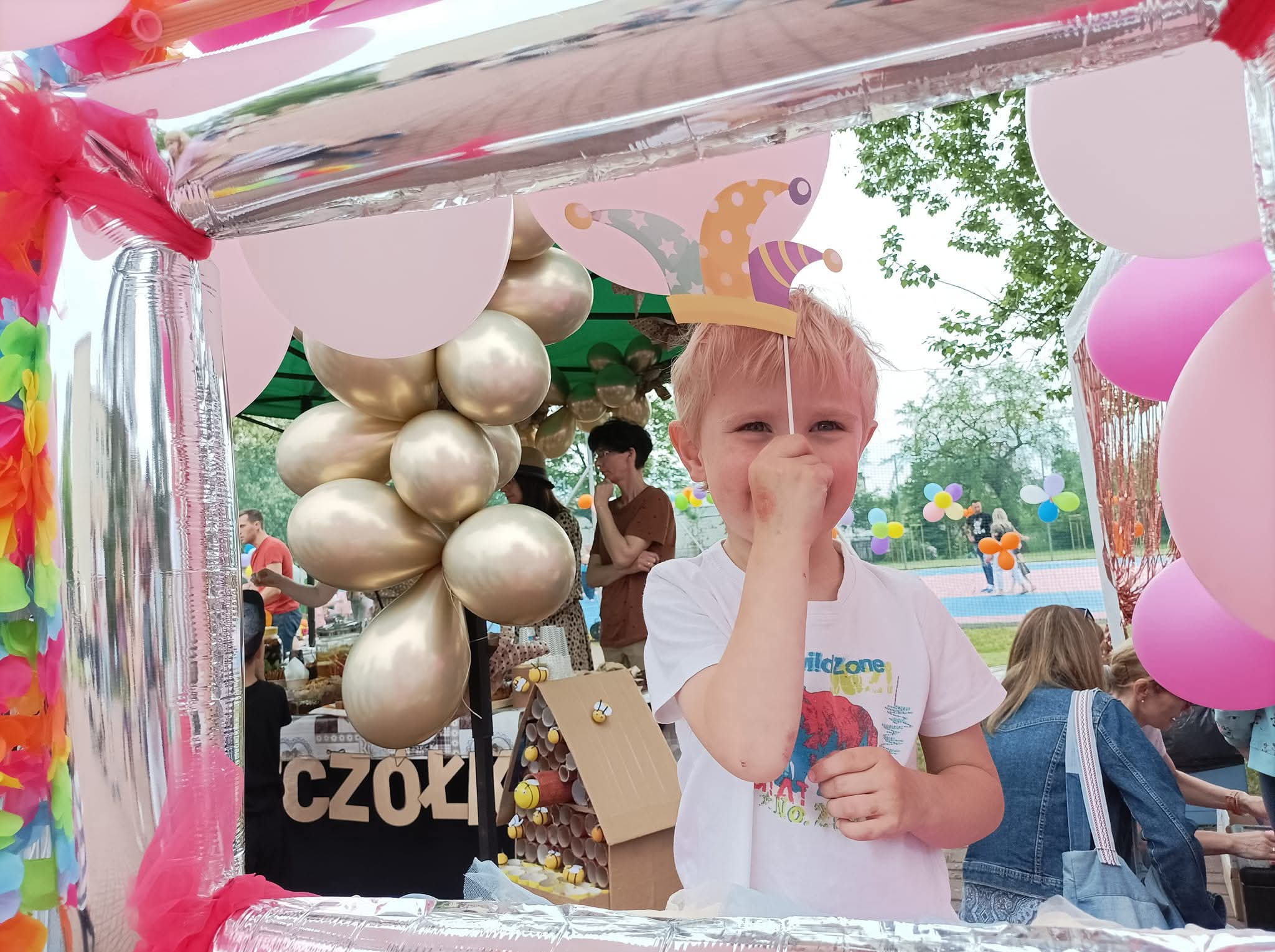 Uśmiechnięty chłopiec z blond włosami trzyma papierową czapkę błazna na tle dekoracji z balonów i srebrnej konstrukcji. Widoczne osoby w tle.