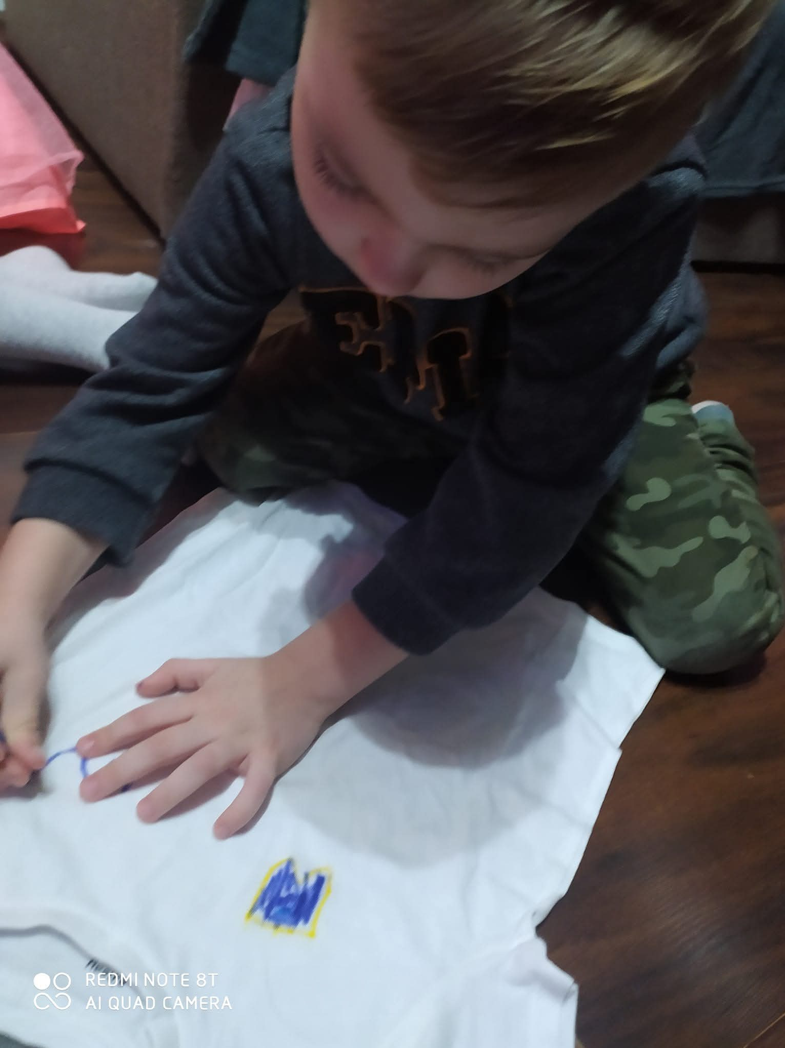 Chłopiec w trakcie malowania wzoru na białej koszulce markerami, siedząc na podłodze. Widoczne dłonie dziecka i fragmenty ubrania. Ujęcie z góry, naturalne oświetlenie.