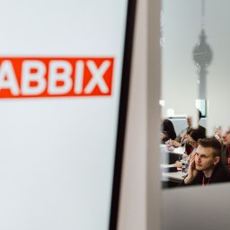 Marcin Krokowski Fotografia Eventowa - Konferencja Zabbix: Uczestnicy skupieni przy stole, widoczni przez szklaną ścianę z logo, w jasnym wnętrzu. Mężczyzna w czarnej koszulce w pierwszym planie.