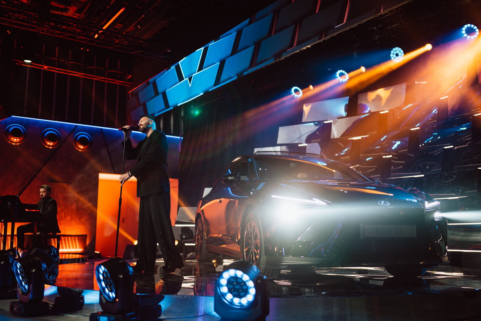 Piosenkarz na scenie z samochodem 'NEW ES' w blasku reflektorów podczas eventu w Warszawie. Pianista w tle, dynamiczne oświetlenie sceniczne.