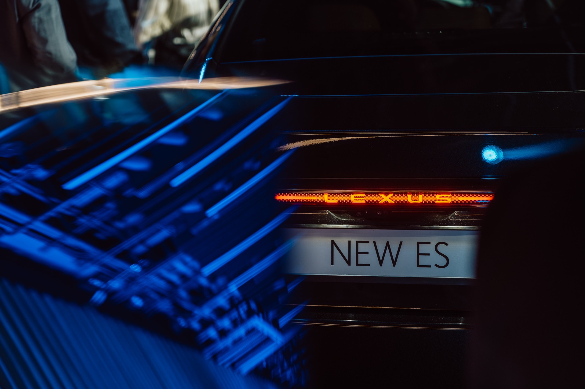 Zbliżenie na tylne światła i tablicę 'NEW ES' czarnego Lexusa, z rozmytym, niebieskim tłem, sugerujące prezentację modelu na evencie motoryzacyjnym.