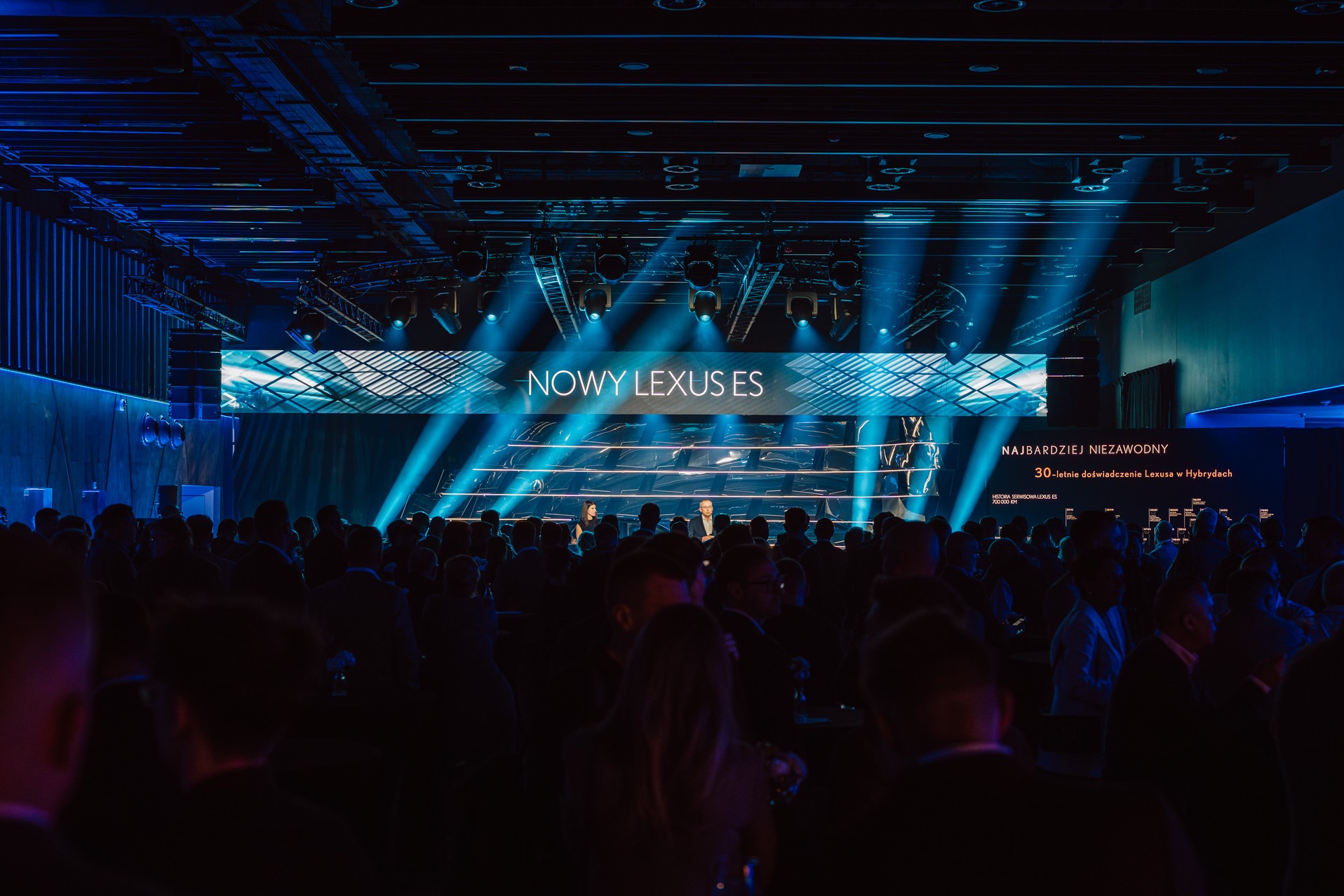 Prezentacja nowego Lexusa ES na evencie. Scena z ekranem, oświetlenie punktowe, tłum gości. Ujęcie z perspektywy widza, dominują ciemne barwy i niebieskie światło.
