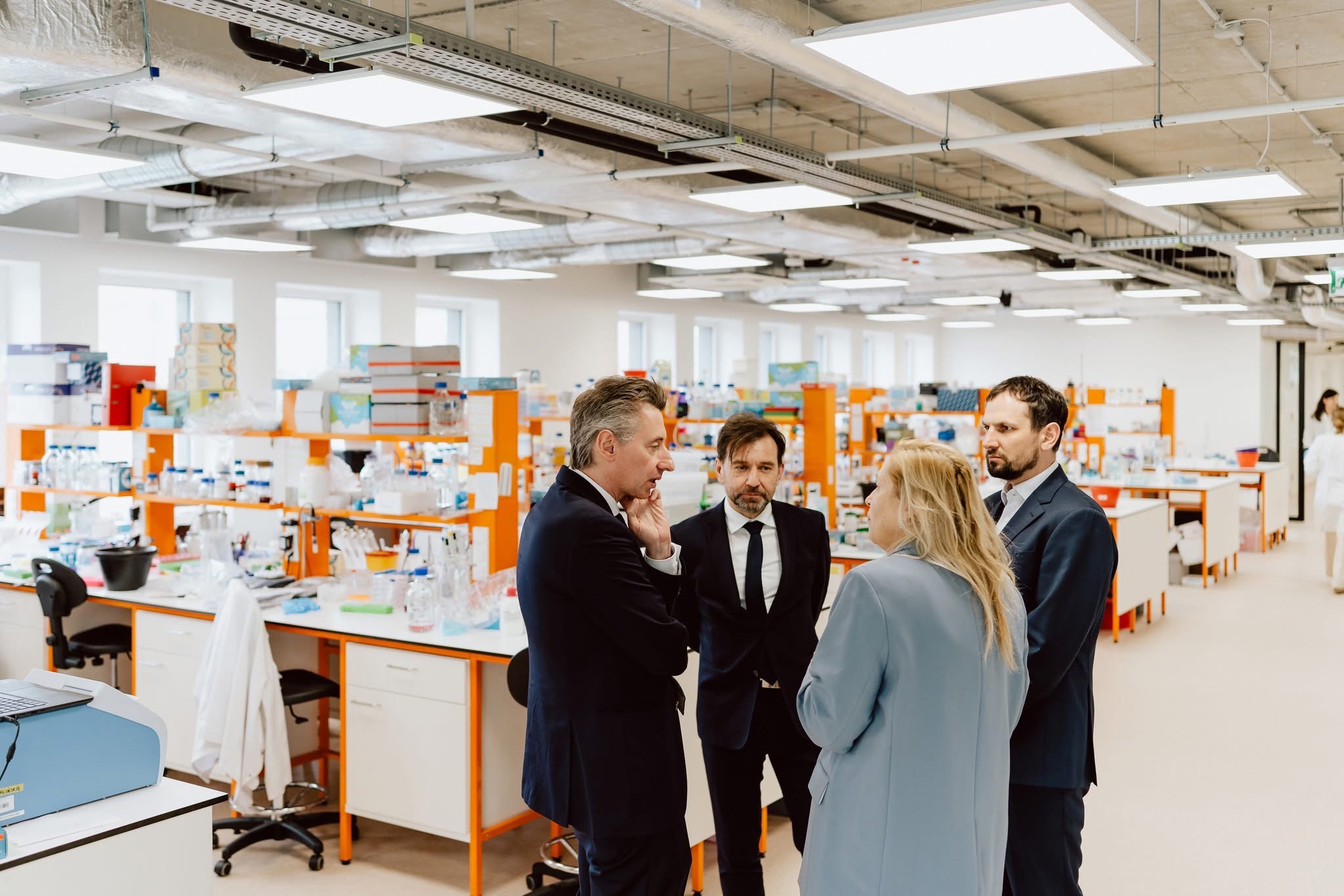 Spotkanie biznesowe w nowoczesnym laboratorium: czterech elegancko ubranych profesjonalistów rozmawia na tle wyposażonych stanowisk pracy.