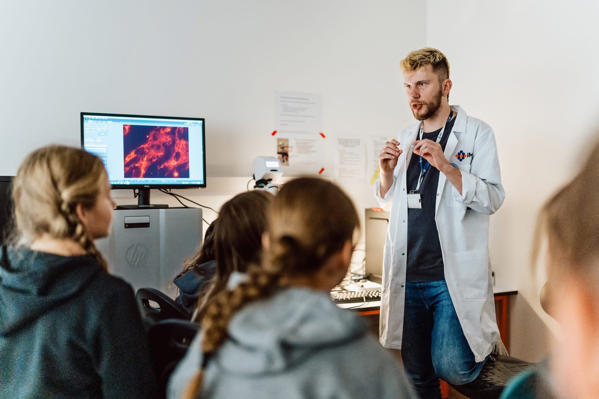 Mężczyzna w białym fartuchu prezentuje wyniki badań na monitorze grupie osób. Widoczne mikroskopowe obrazy komórek. Jasne wnętrze laboratorium.