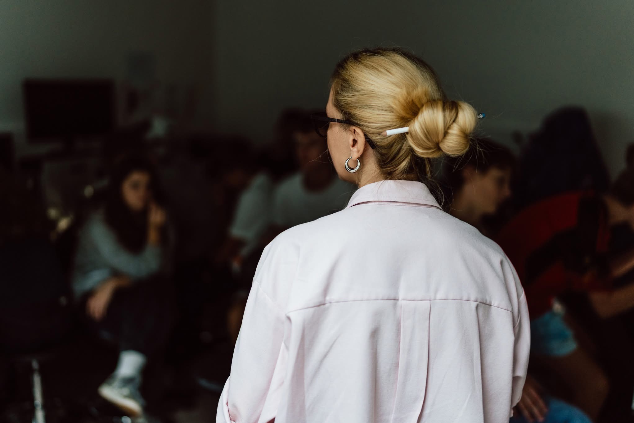 Blondwłosa kobieta w okularach, odwrócona tyłem, z upiętymi włosami i ołówkiem, w jasnoróżowej koszuli, na tle rozmytej grupy osób w pomieszczeniu.