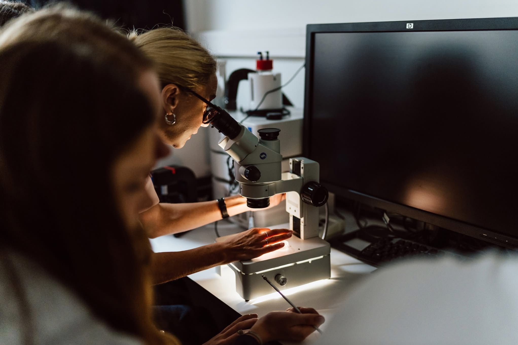 Praca w laboratorium w Warszawie: kobieta w okularach patrzy przez mikroskop, obok monitor i klawiatura. Precyzyjne badania pod lupą.